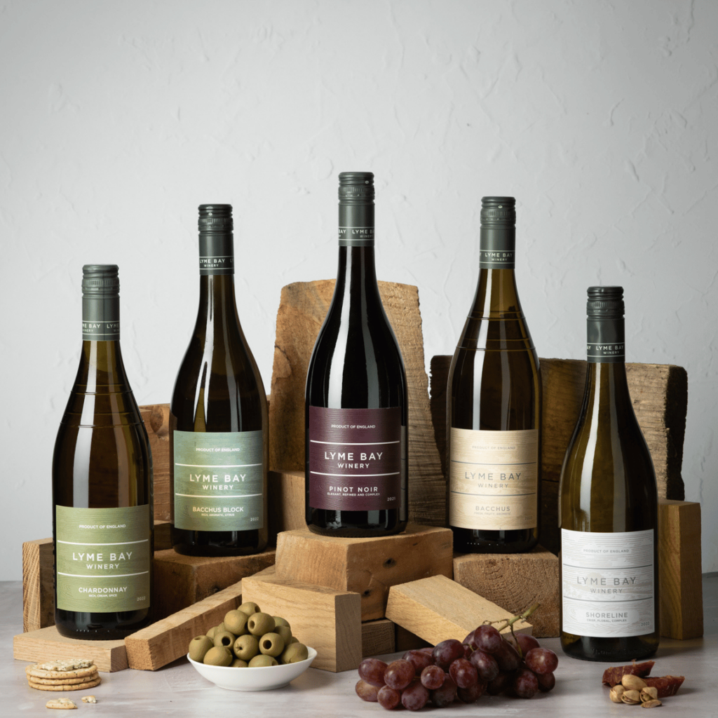 Five bottles of Lyme Bay Winery wine displayed on wooden blocks, with a bowl of green olives, crackers, red grapes, and nuts arranged in the foreground against a light background.